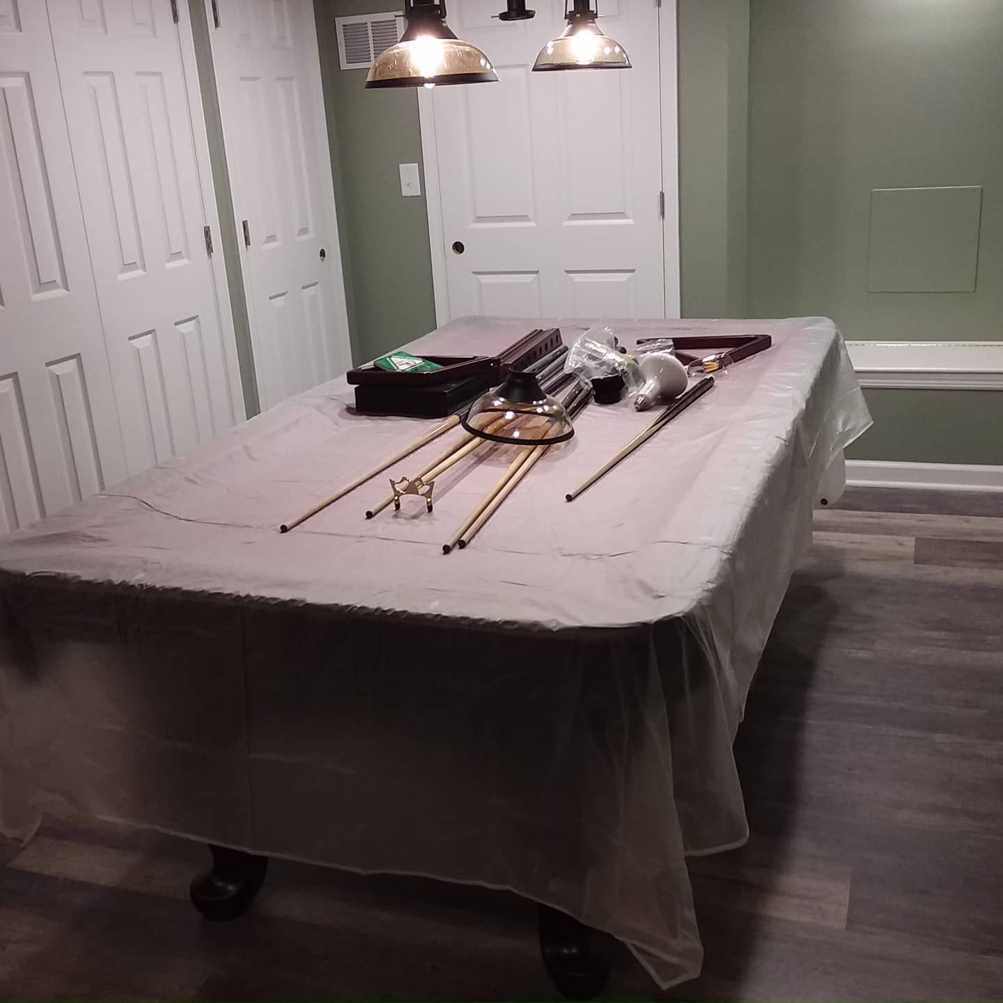 Pool table covered with a white sheet, pool cues and accessories on top, in a room with white doors.