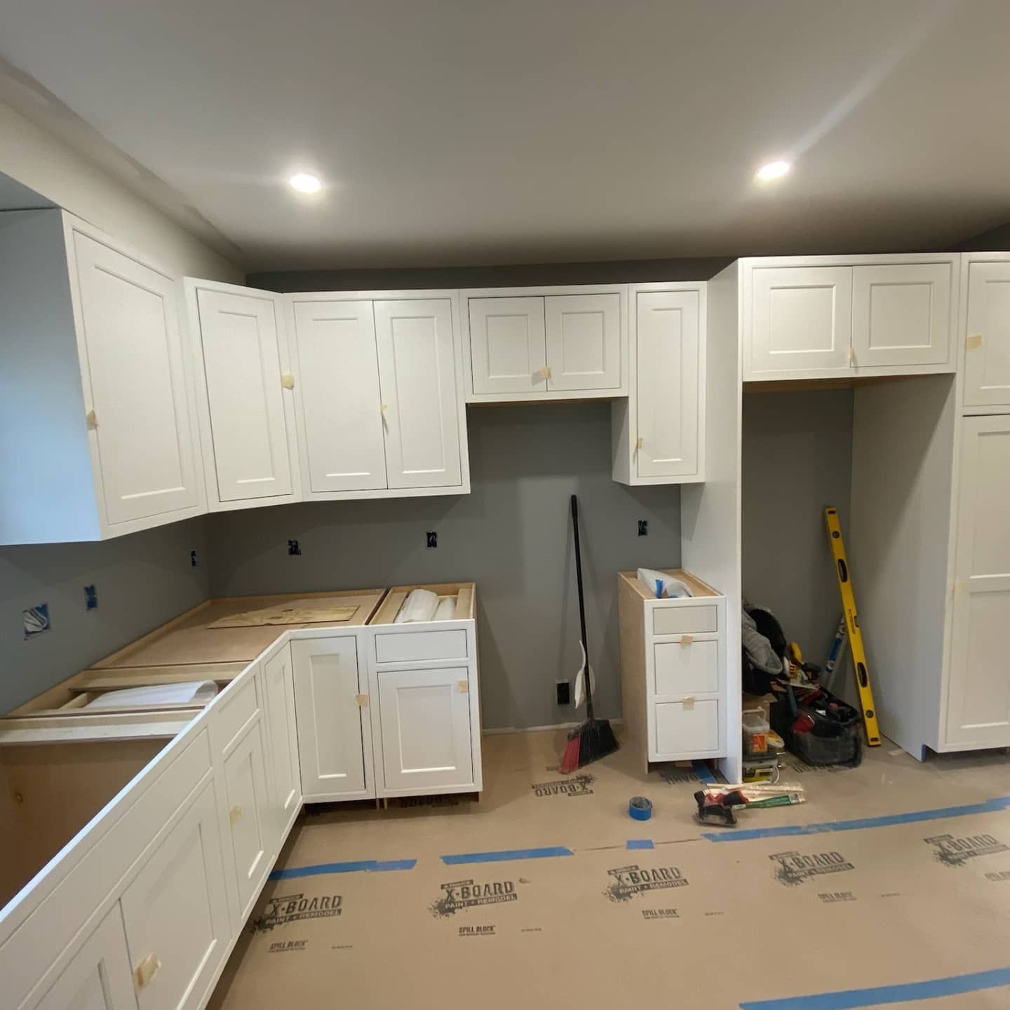 Kitchen cabinets installation in progress; white cabinets, gray walls, light flooring, tools and supplies visible.