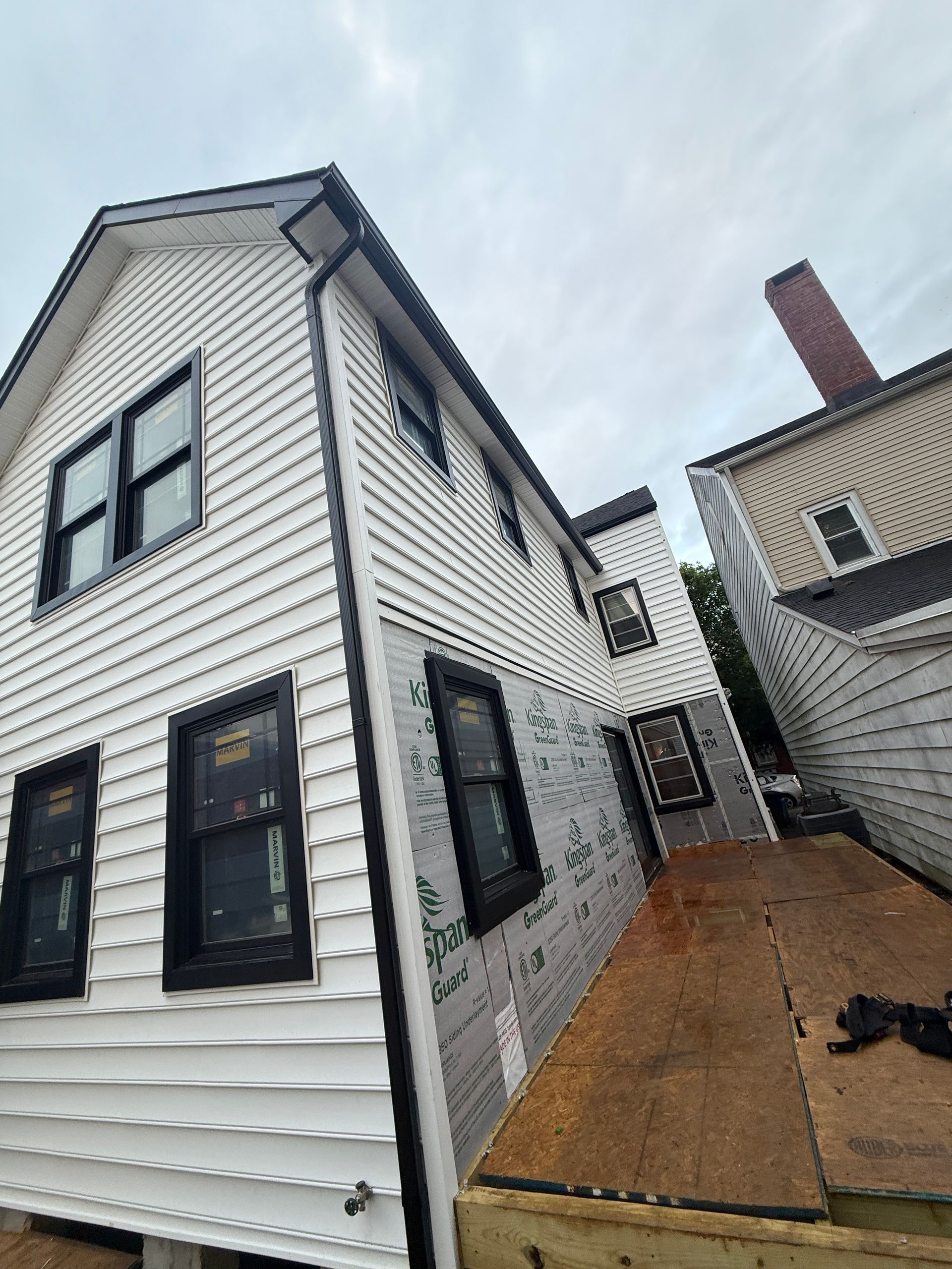 Exterior view of a two-story house under construction. White siding, black window frames, adjacent building with chimney.