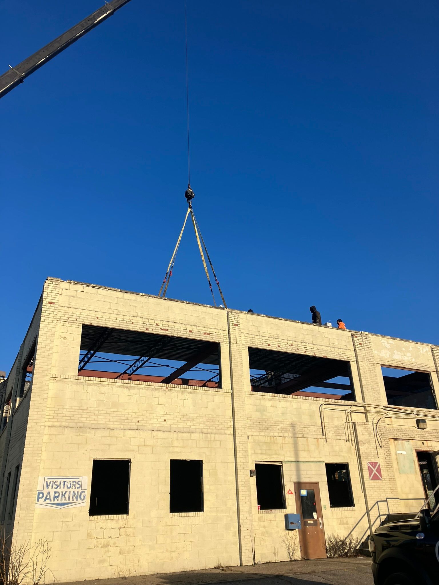 Crane lifting a section of a building's roof under a bright blue sky. Construction workers are visible.