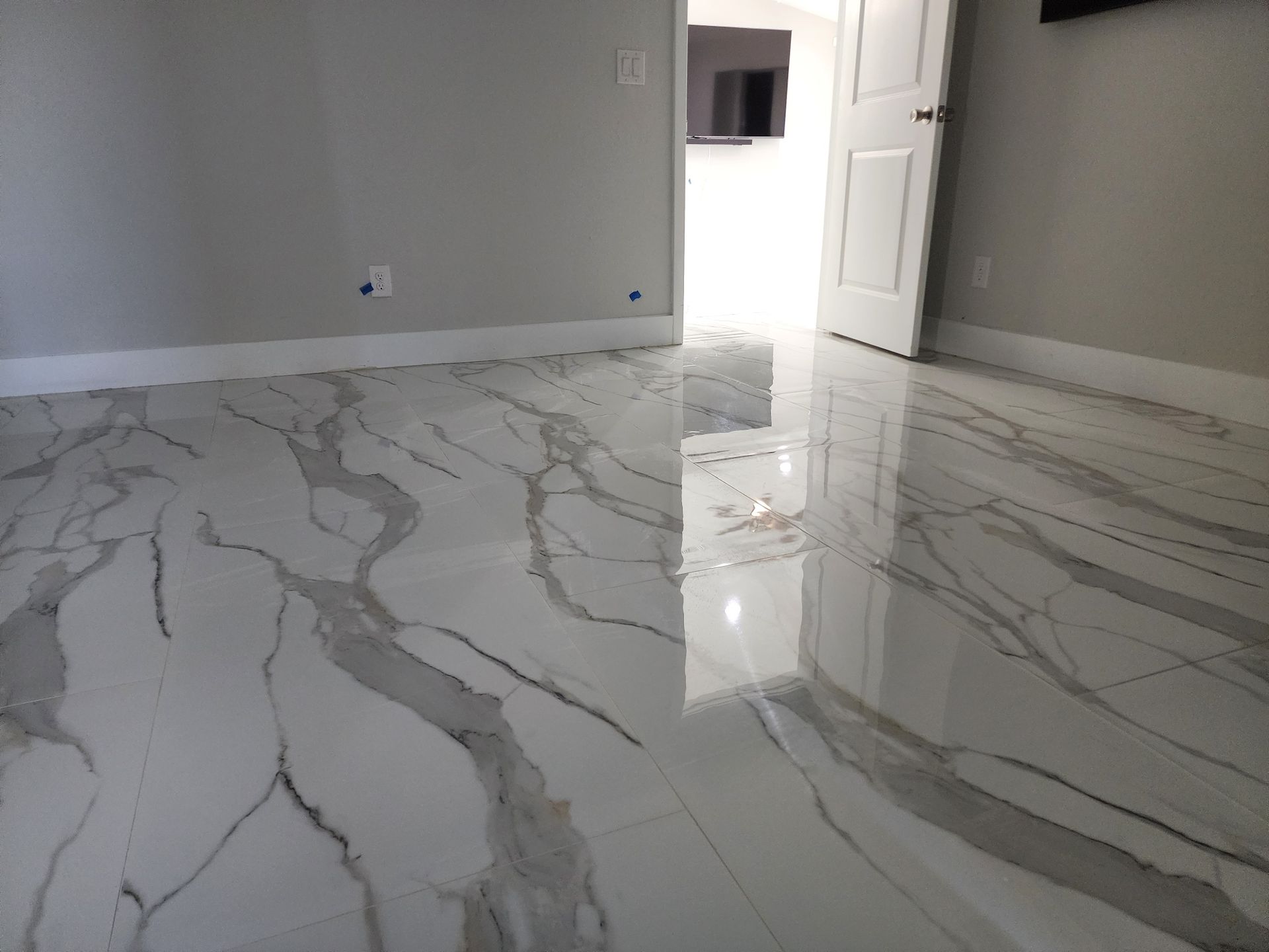 White and gray marble-look floor in a room with light gray walls, open white door, and a glimpse of a TV.