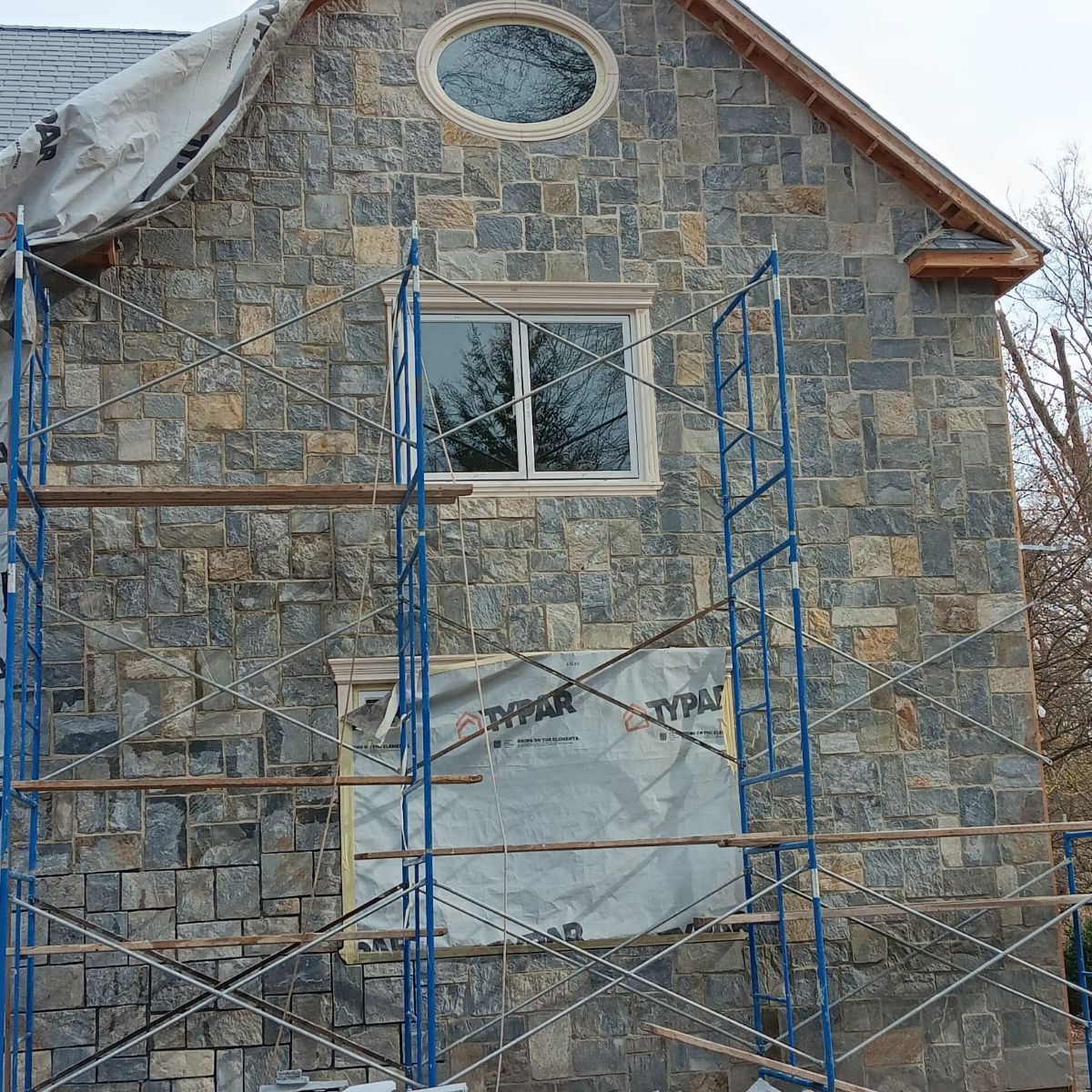 Stone-clad house exterior under construction; scaffolding surrounds a window; Tyvek wrap visible.