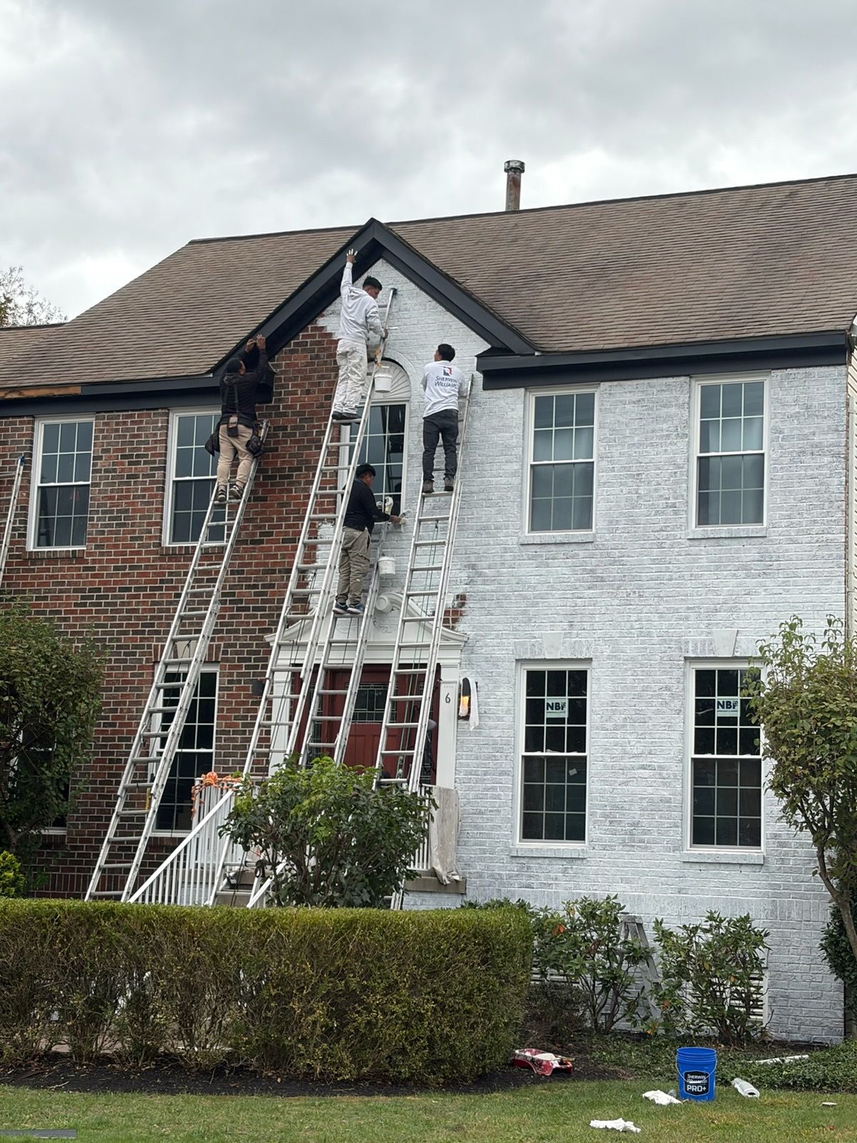 People painting a two-story brick house white, using ladders. Overcast sky.