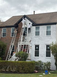 People painting a two-story brick house white, using ladders. Overcast sky.