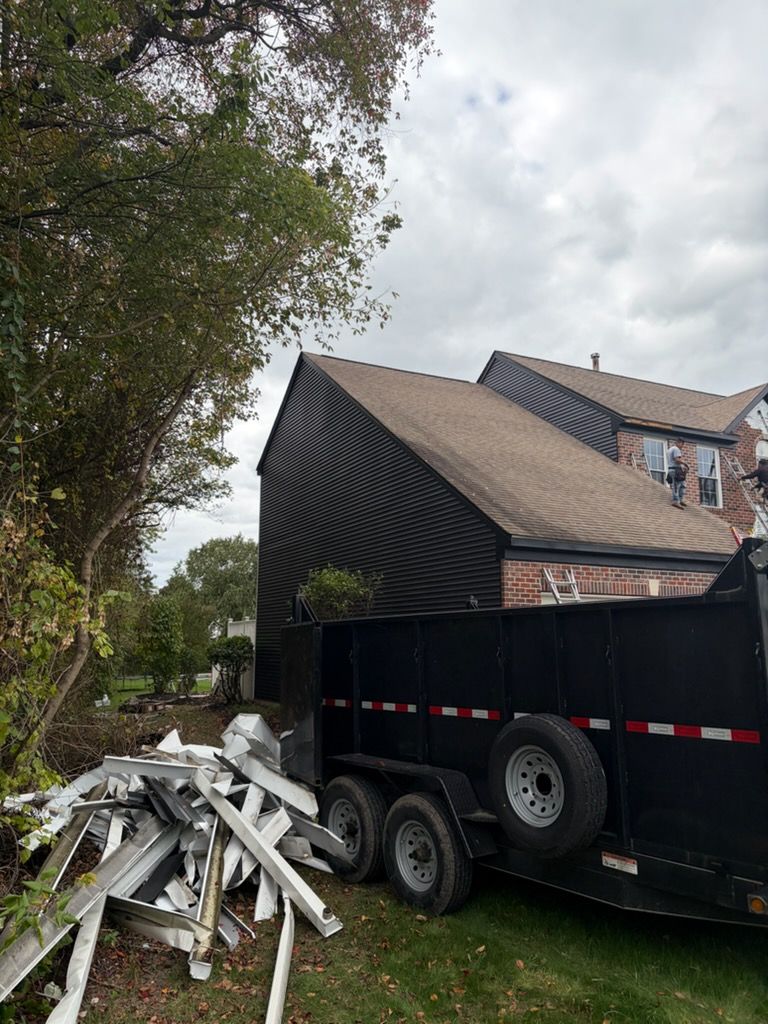 A black dump trailer next to a house with debris and tree in the background. Overcast sky.