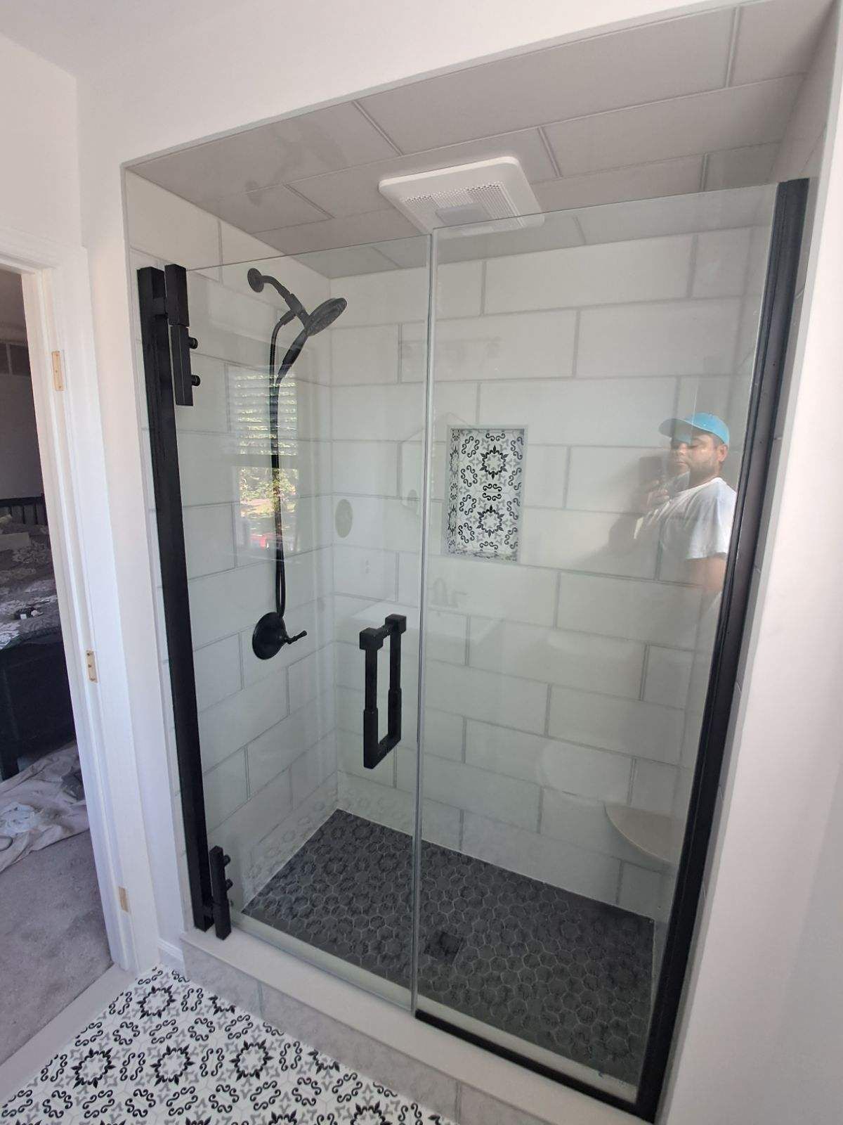 Black-framed glass shower with white tile walls, a patterned floor, and a person reflected in the glass.