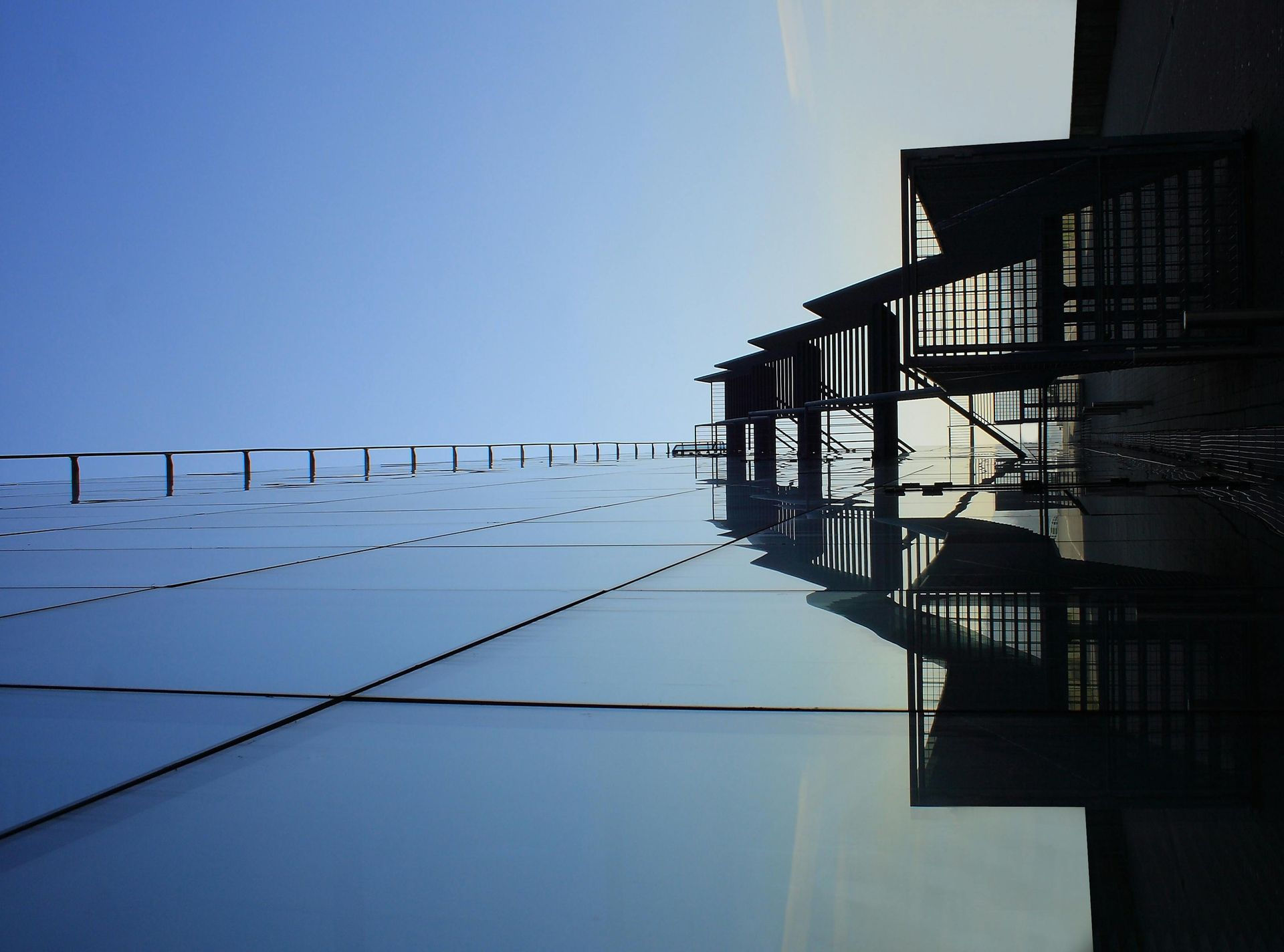 Building reflected in a large glass surface against a blue sky. Sunlight casts shadows.
