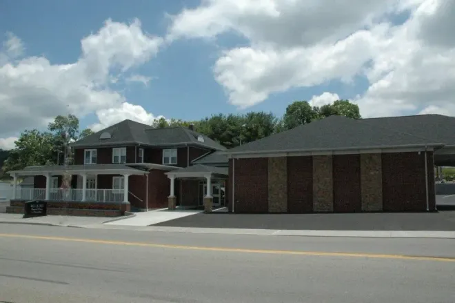 Two-story brick building with a porch and flat-roofed brick extension on a sunny day with a cloudy sky.