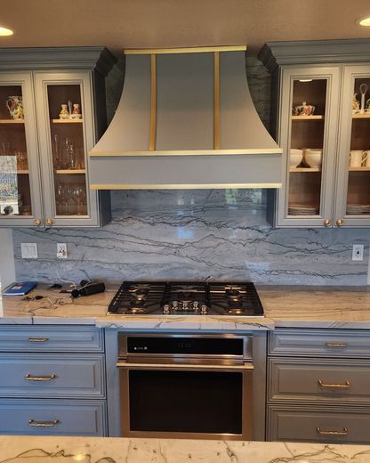 Blue kitchen with a grey stove hood over a cooktop and cabinets with gold hardware.