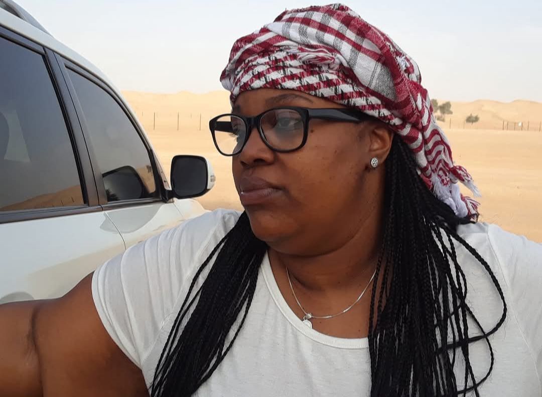 A woman wearing glasses and a head scarf is standing in front of a car.