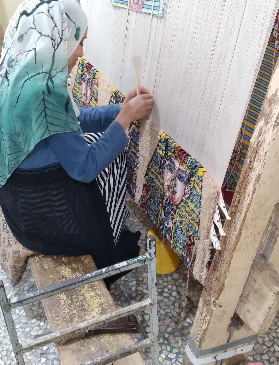 A woman is sitting on a chair making a rug on a loom.