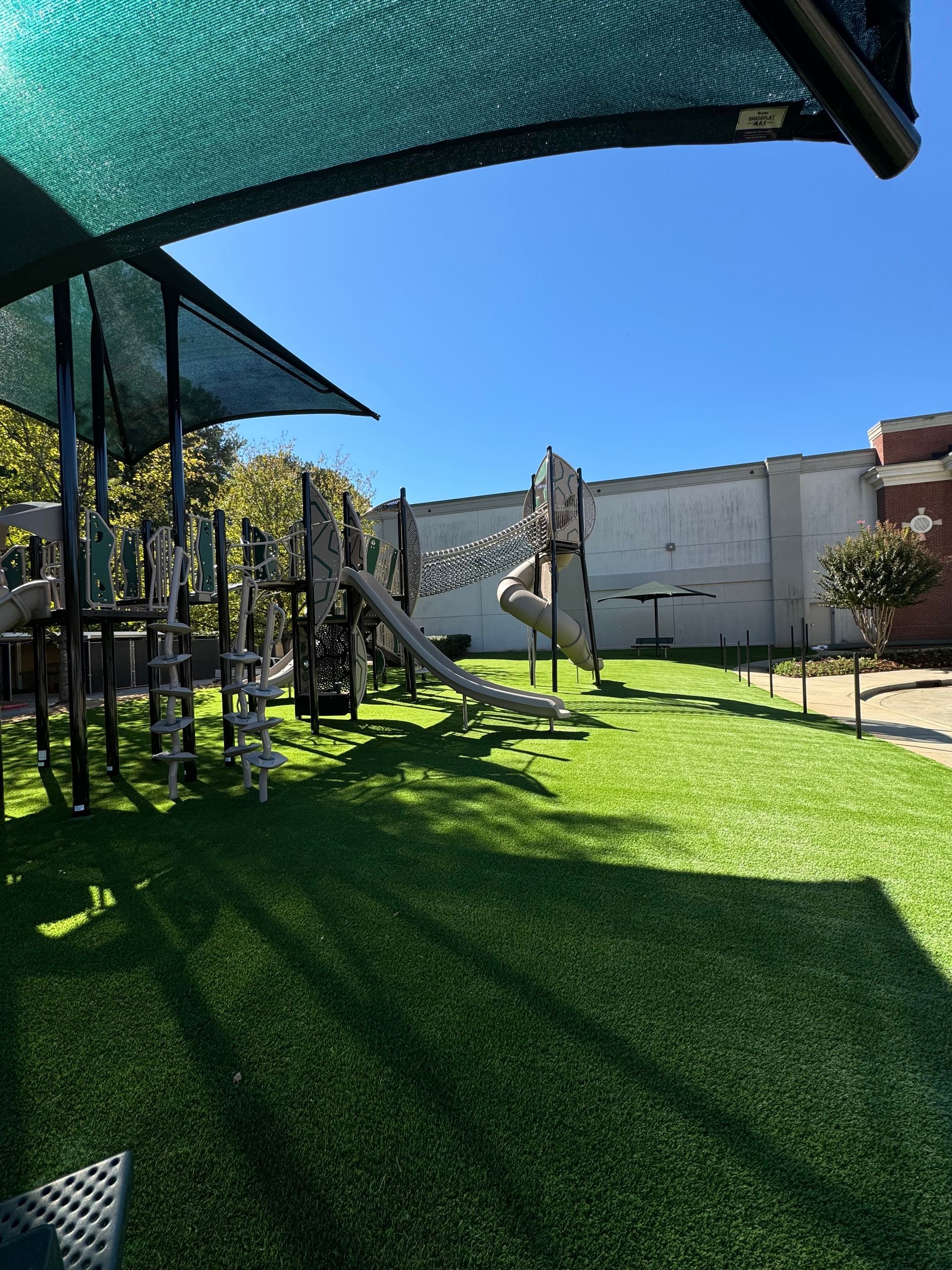 Playground with green turf, equipment under a shade, against a white wall and blue sky.