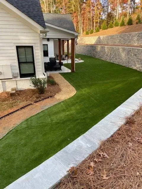 A house with white siding and green turf. A curved pathway leads to a porch with seating. A retaining wall borders the yard.
