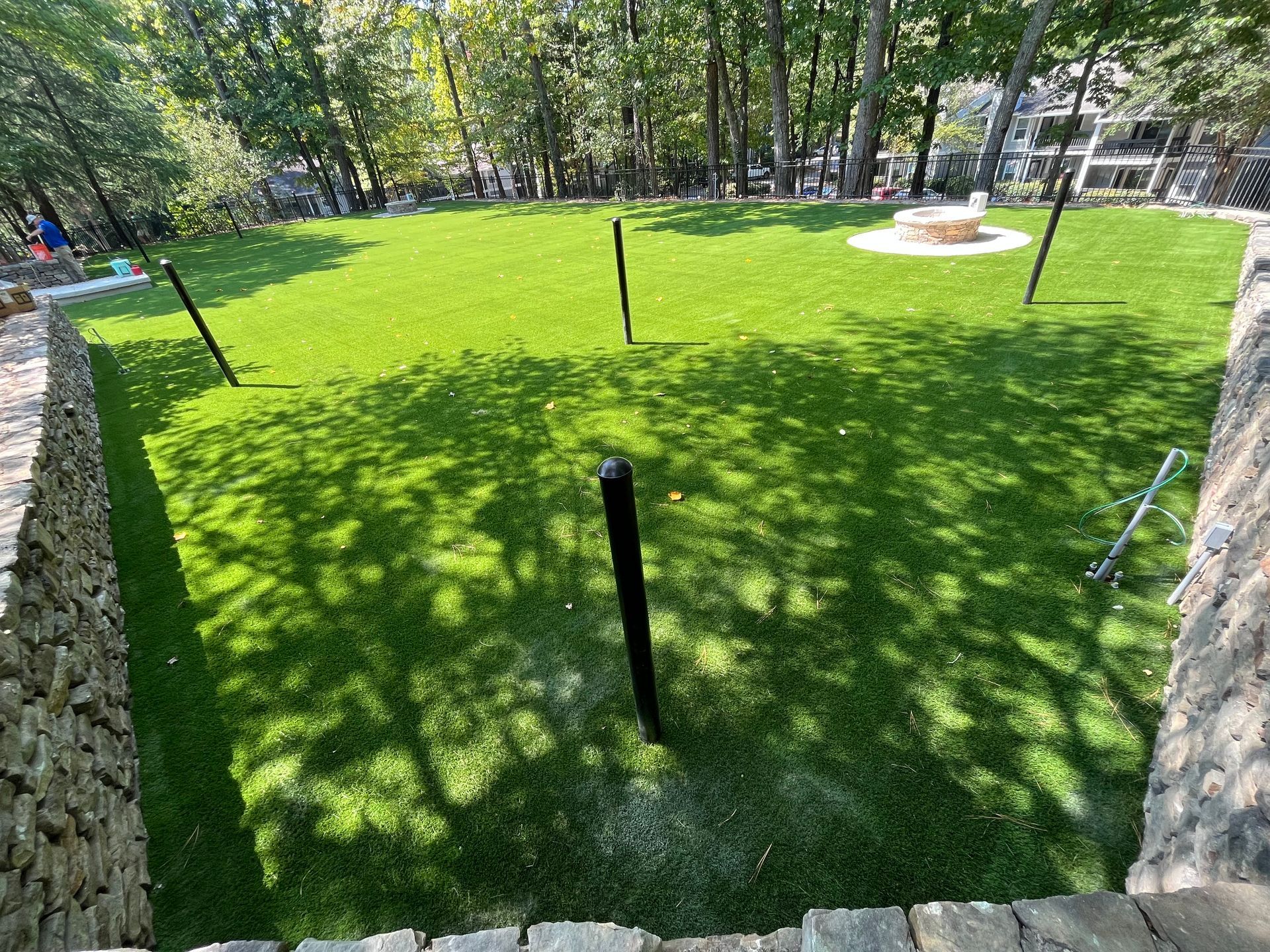 Green lawn in a yard with black poles, surrounded by stone walls and trees; a fire pit is in the background.