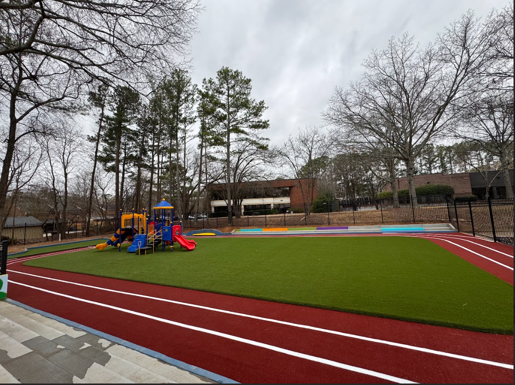 Playground with running track, green turf, colorful equipment, and surrounding trees under a cloudy sky.