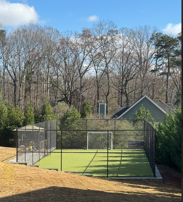 A fenced, artificial turf soccer area in a backyard, with a goal, next to trees and a house.