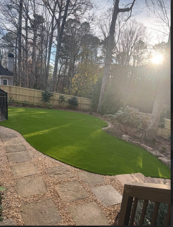 Green lawn in backyard, stone path, wooden fence, trees, and bright sunlight.