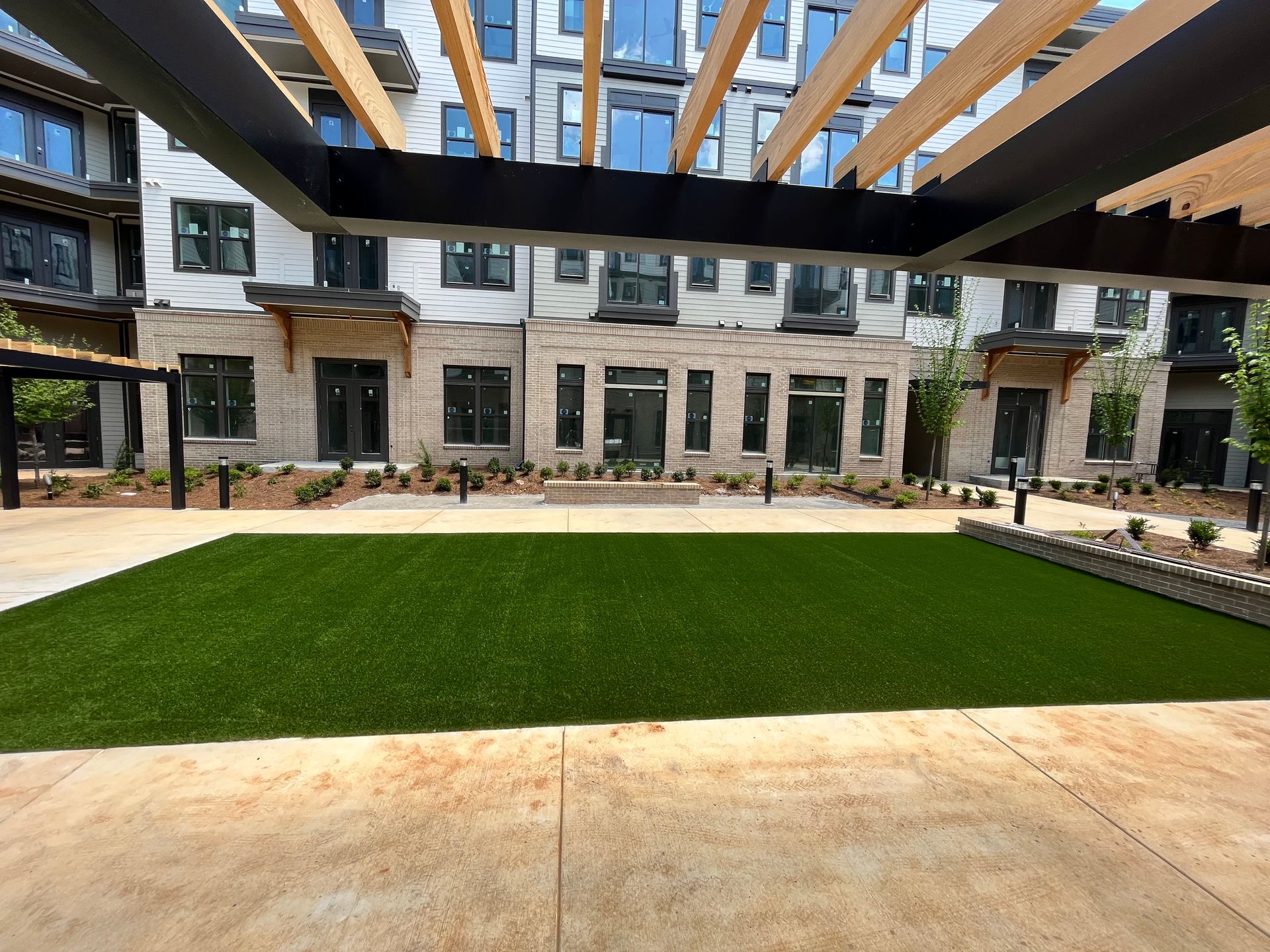 Courtyard with artificial turf, seating area, and building with windows under a wooden trellis.