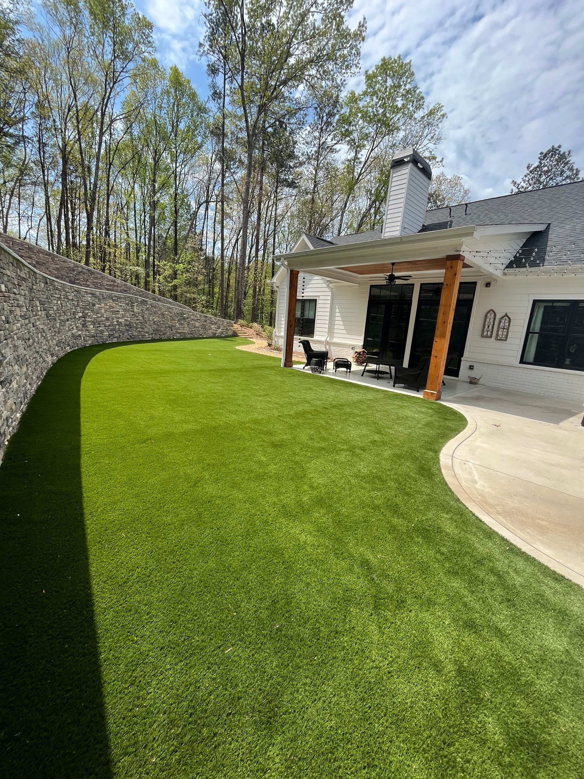 Green lawn curves around a white house with a covered patio; trees and blue sky background.