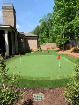 A putting green surrounded by a stone wall, trees, and lush green grass in a backyard setting.