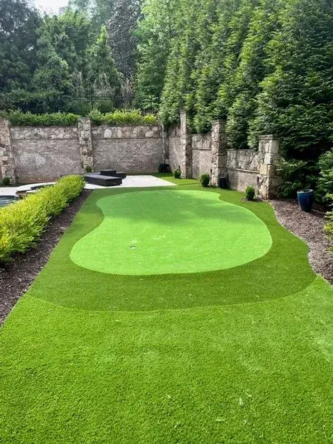 Artificial putting green surrounded by lush green lawn and stone wall in a backyard setting.