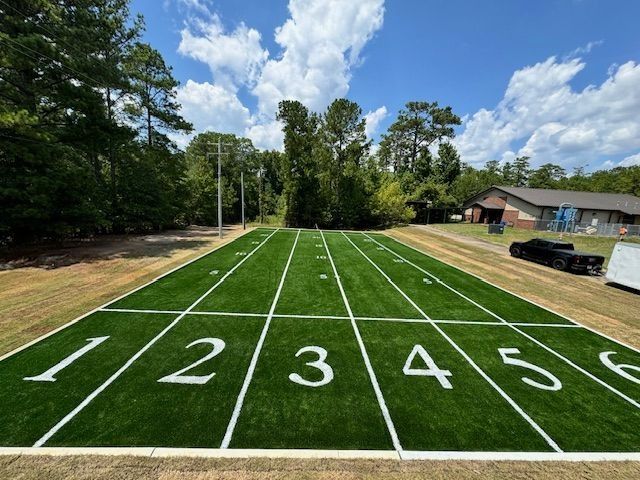Green turf field with white yard lines and numbered sections; trees and a building in the background.