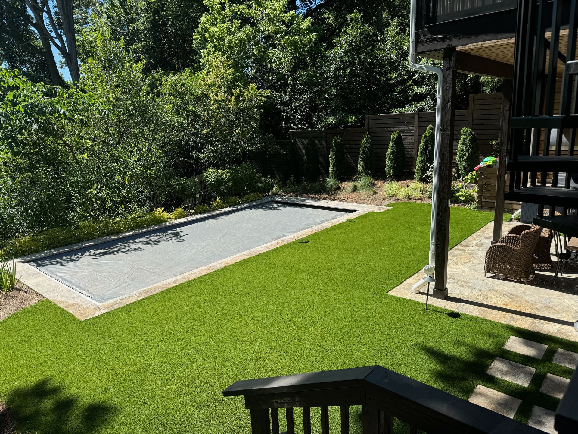 Green lawn surrounds a rectangular pool with a closed cover, adjacent to a wooden deck and lush trees.