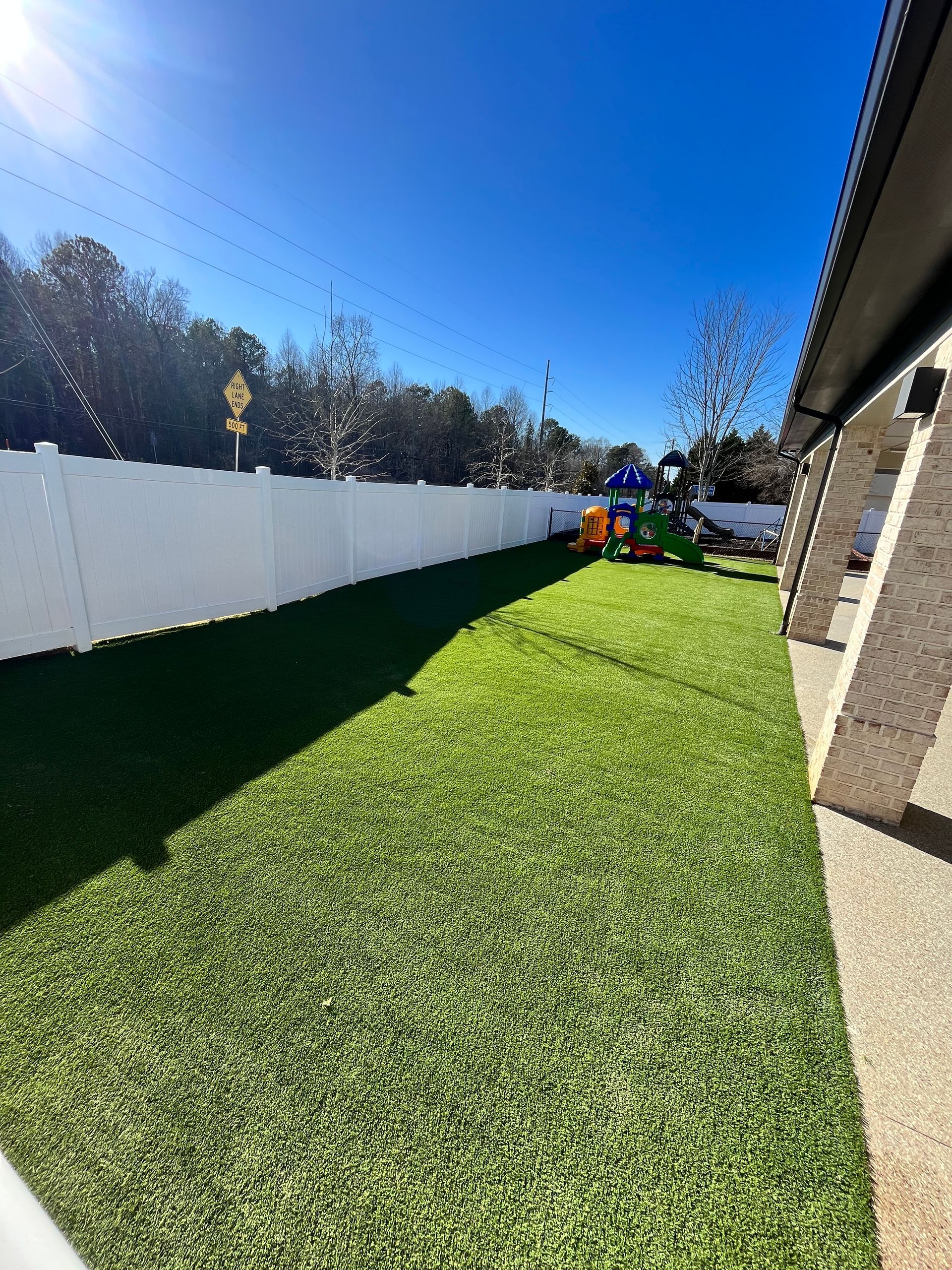 Green turf backyard with white fence, playground, and blue sky.