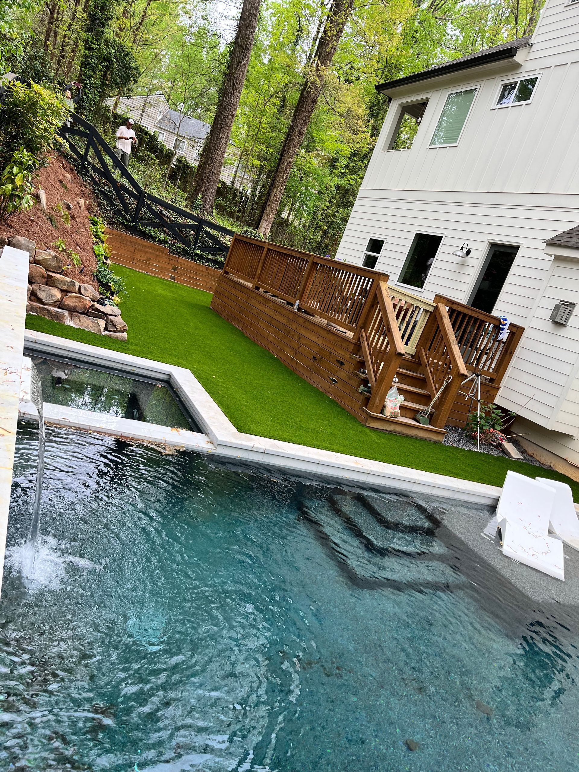 Pool with waterfall, wooden deck, green turf, and white house on a sloped yard.
