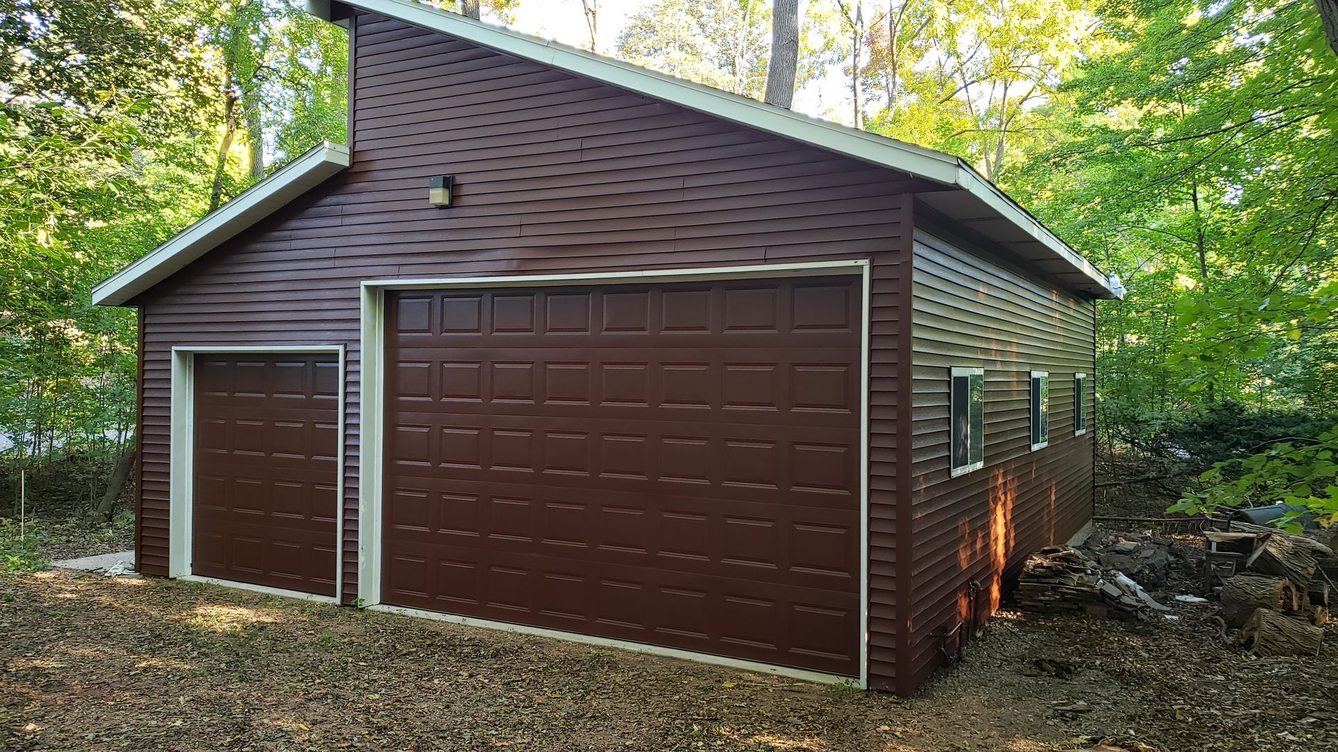 A brown garage with two garage doors is sitting in the middle of a forest.