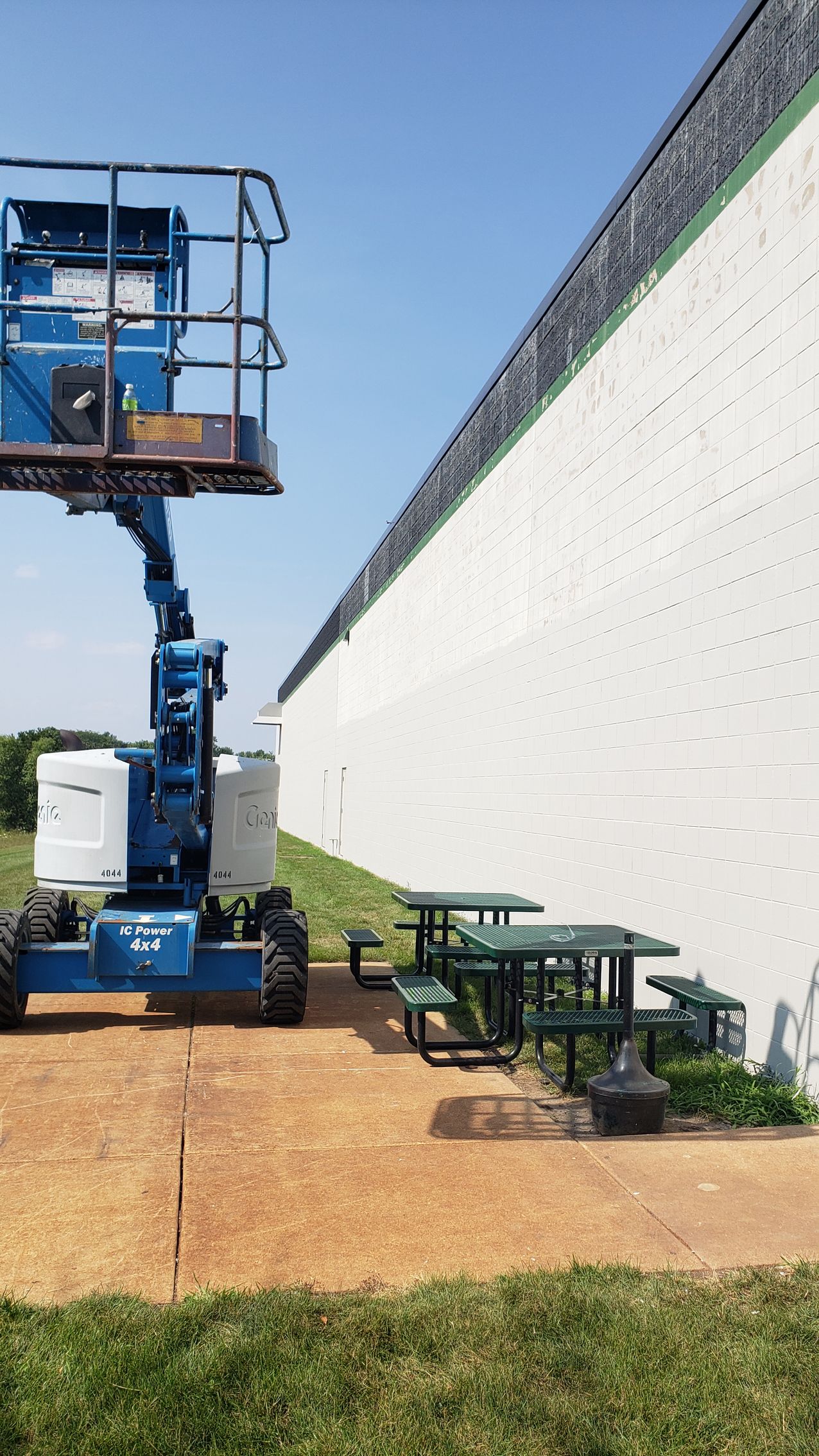 A blue aerial lift is sitting in front of a white building next to a picnic table.