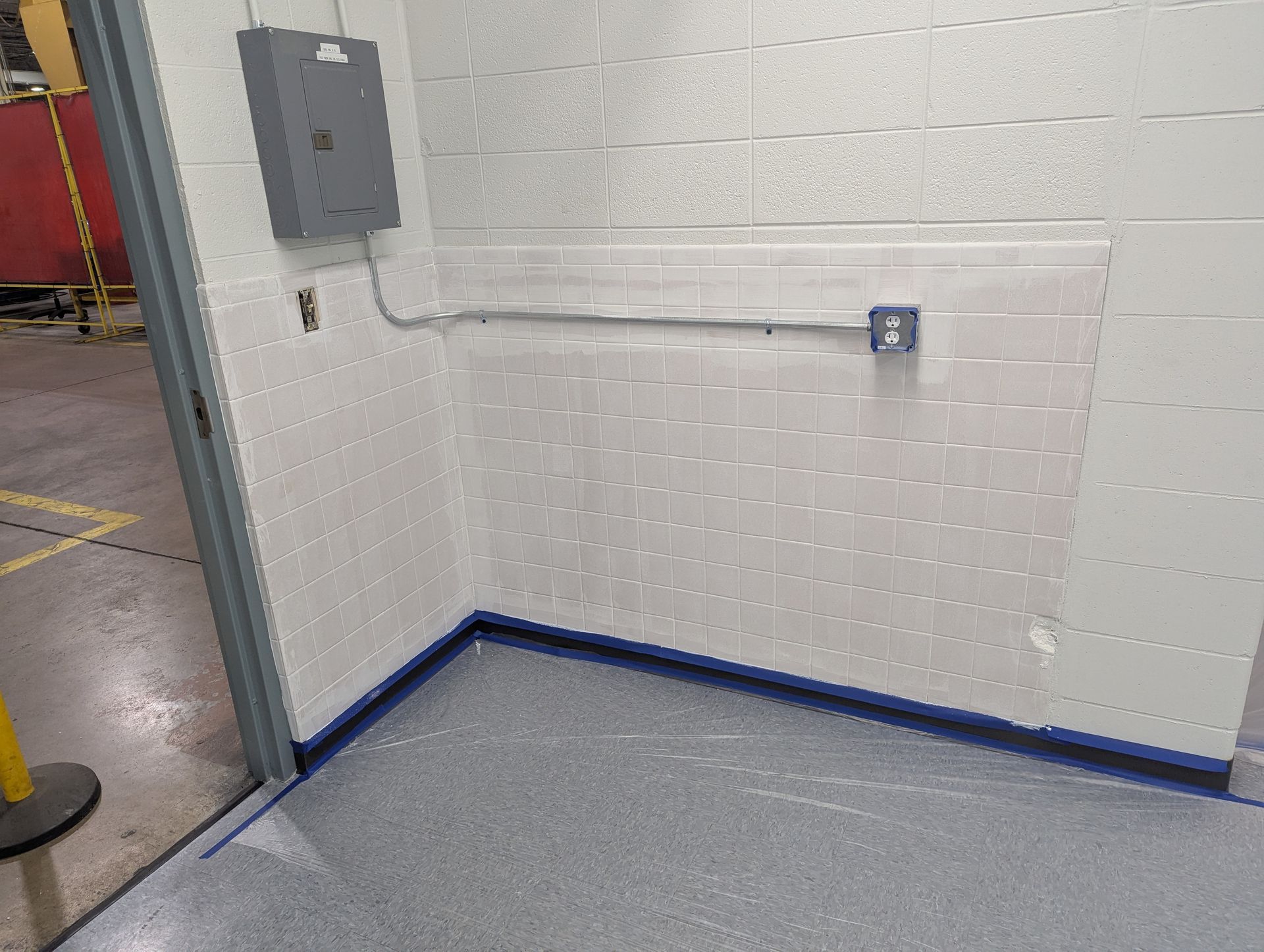 Corner of a room with gray floor, white tiled walls, electrical box and outlet.