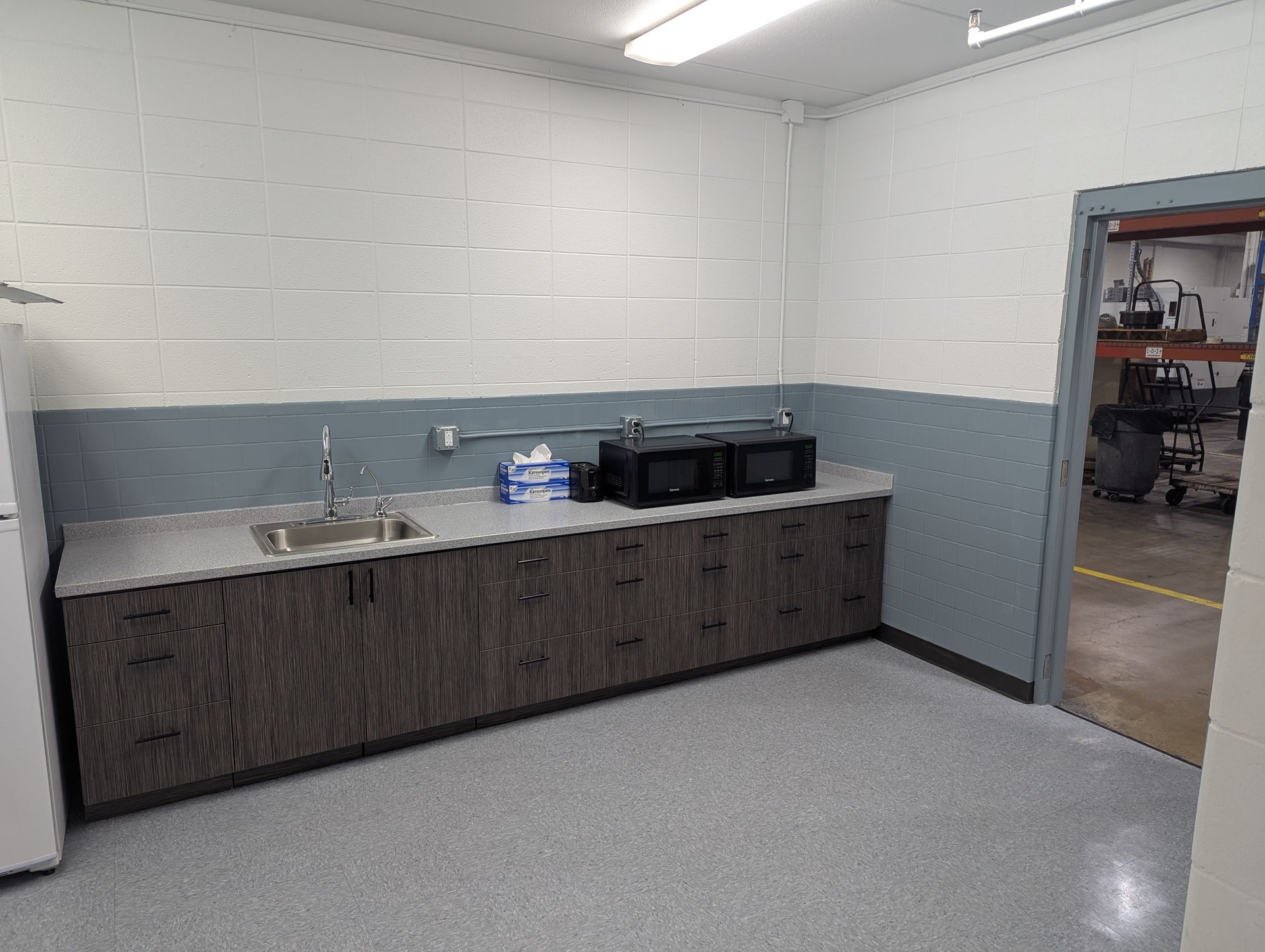 A small breakroom with a sink, cabinets, microwaves, and a refrigerator, and a doorway leading to another room.