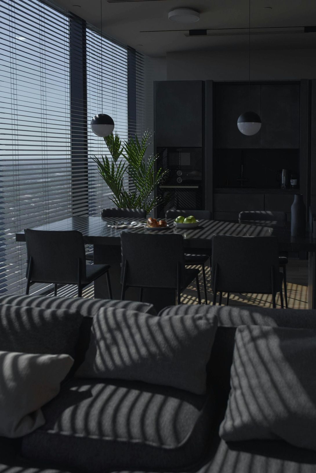 Sala de jantar escura, luz do sol através das persianas. Mesa posta com frutas, cadeiras ao redor. Sofá cinza em primeiro plano.