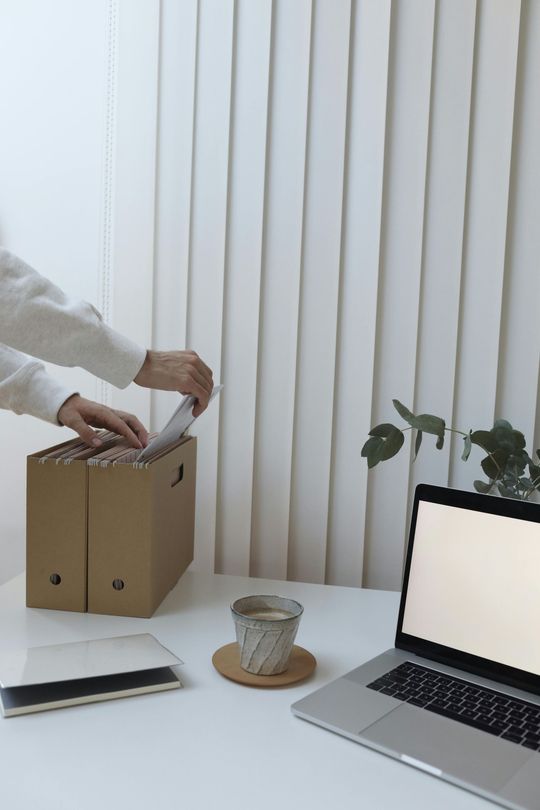 Uma pessoa está colocando papéis em uma caixa de papelão ao lado de um laptop, no fundo uma persiana