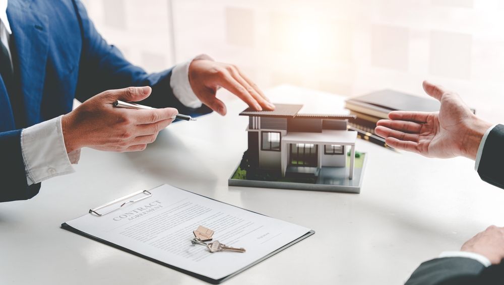 Two people at a table, discussing a house model, contract, and keys. One person gestures toward the model.
