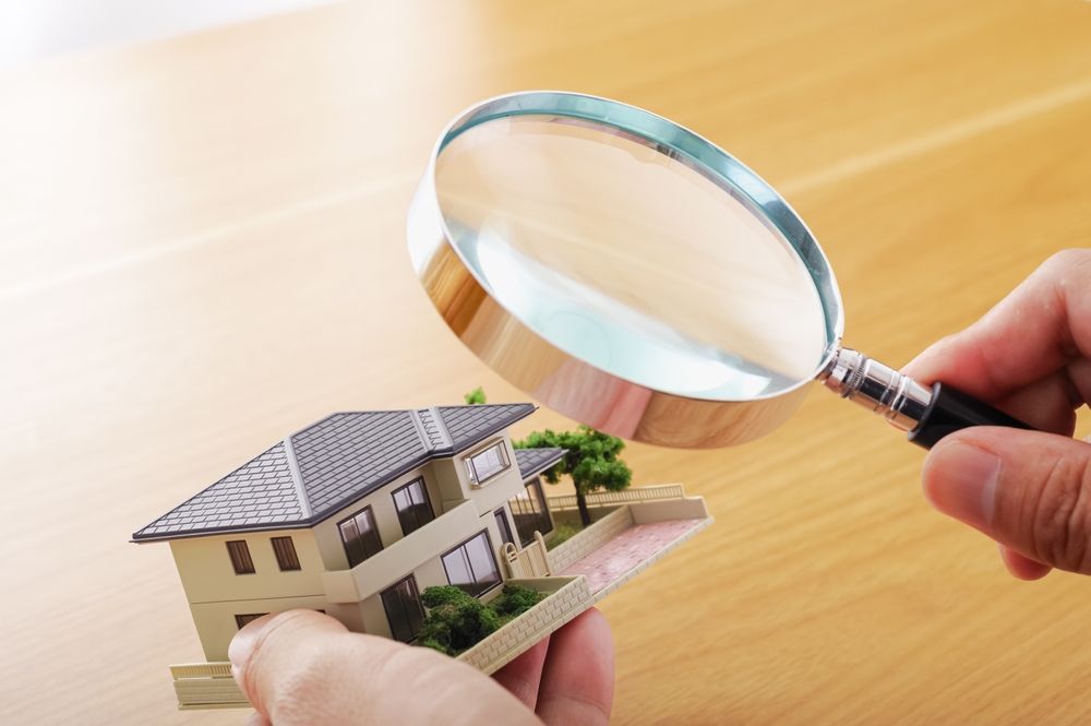 Hand holding magnifying glass over miniature house model on wooden table.