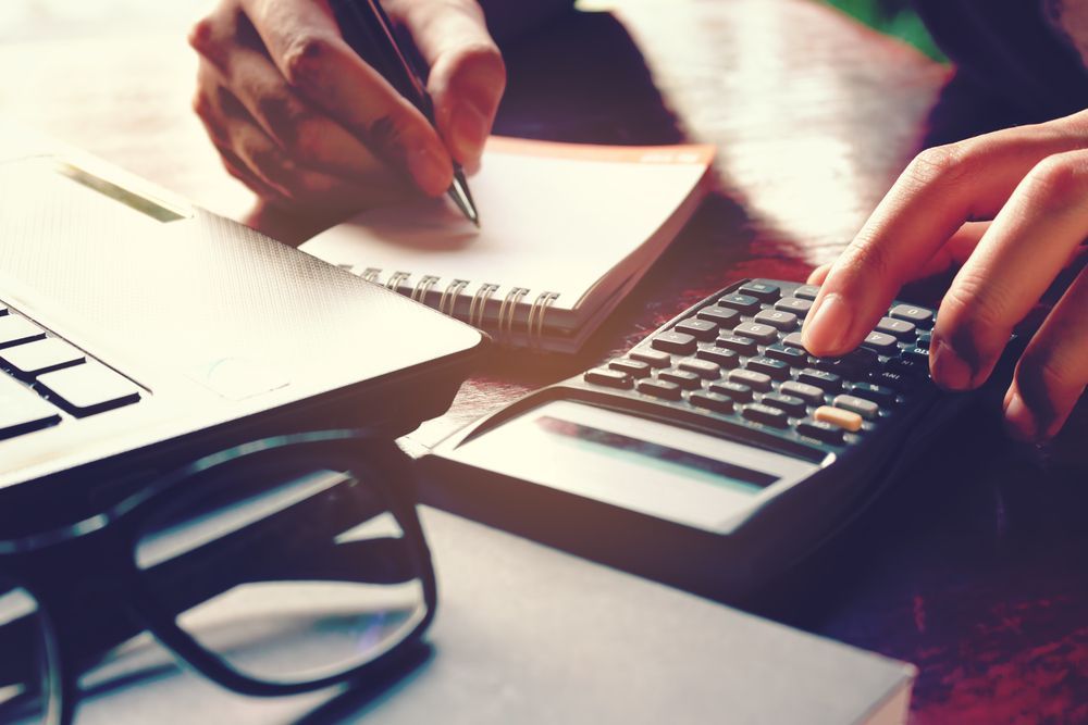 Person using a calculator, taking notes, with laptop and glasses on a desk.