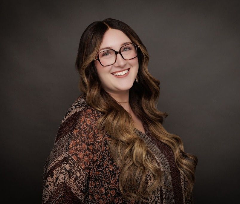 Emily Sibley, LCSW with glasses, smiles, wearing a patterned cardigan against a gray backdrop.