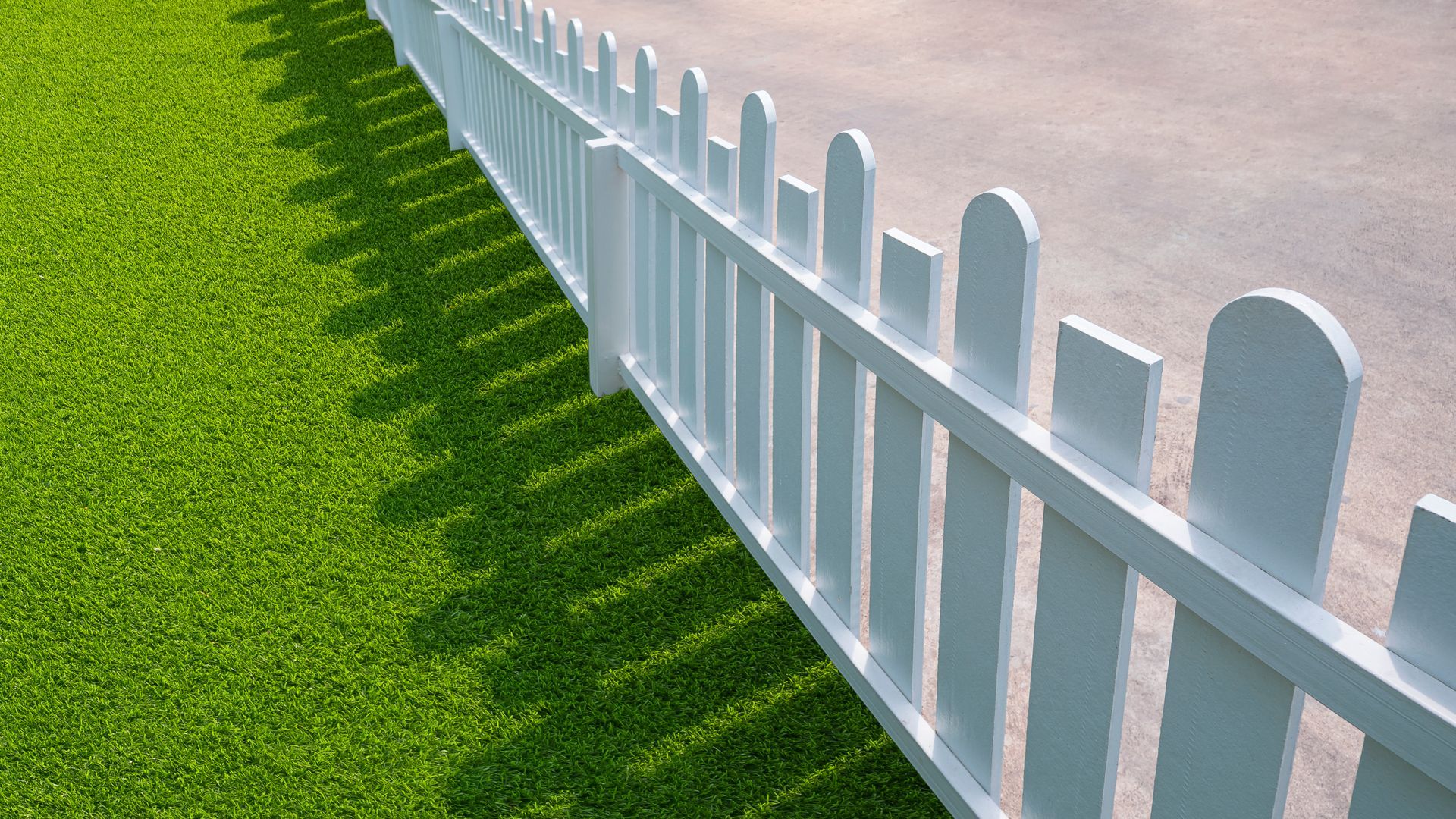 White picket fence casting a shadow on bright green grass and a gray asphalt surface.