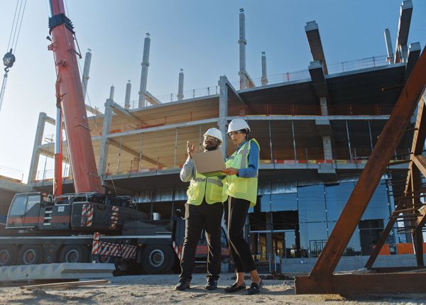 Two construction workers, a man and a woman, in hard hats and vests review plans on a laptop at a construction site.