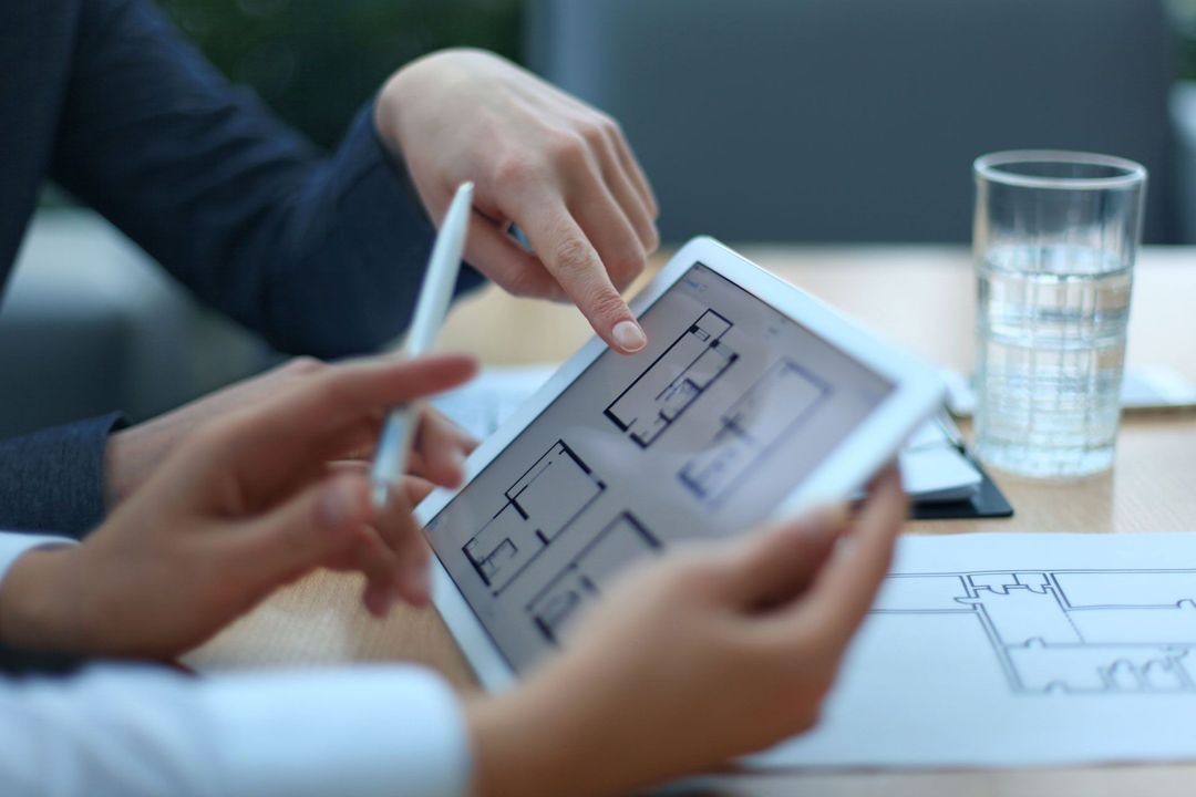 Two people pointing at a digital floor plan on a tablet at a desk, reviewing architectural blueprints.