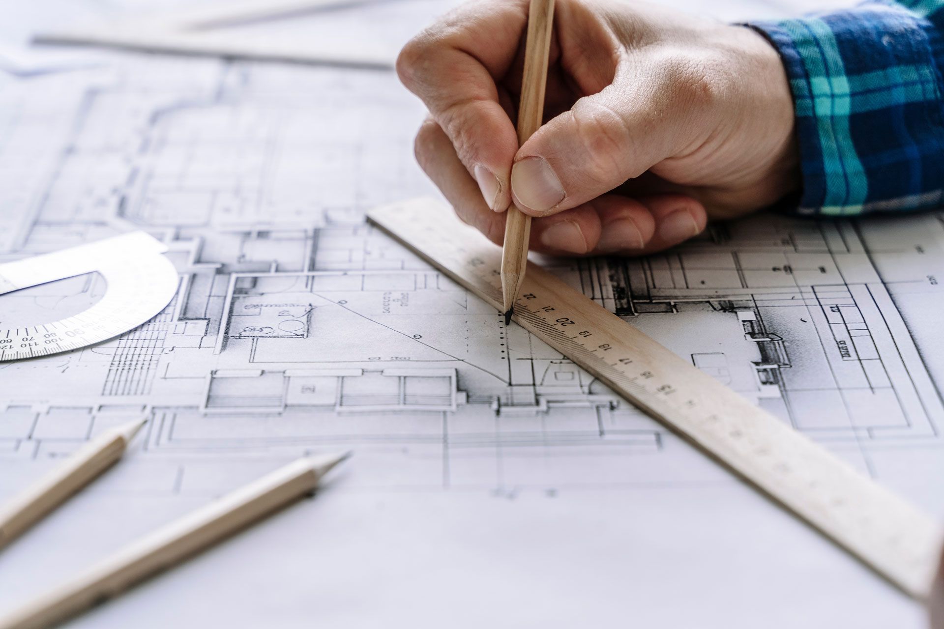 A person's hand sketching architectural plans with a pencil and ruler on a blueprint-filled desk.