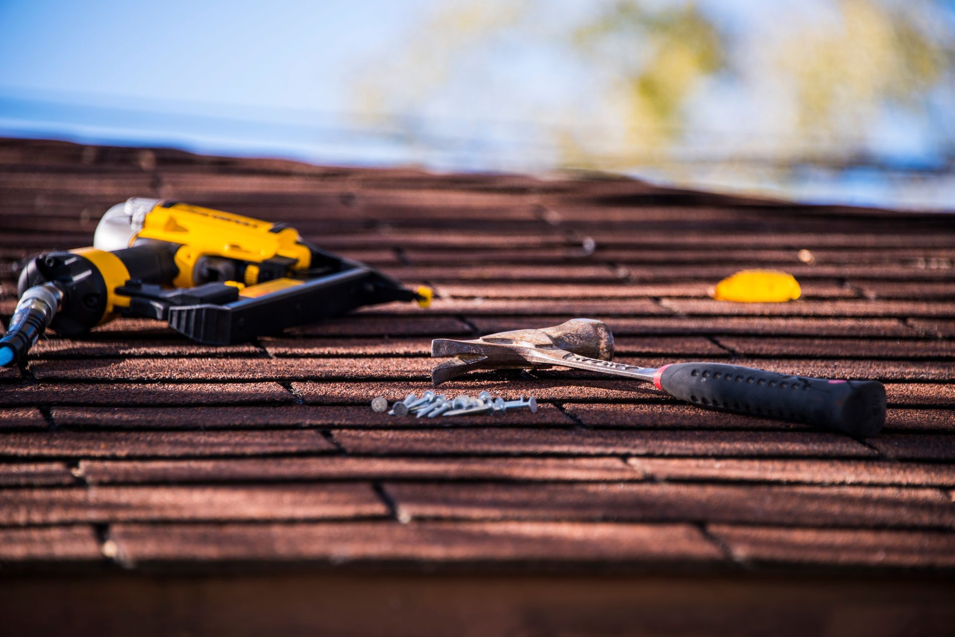 Roofing tools on a shingled roof, including a yellow drill and hammer, with blue sky in the background