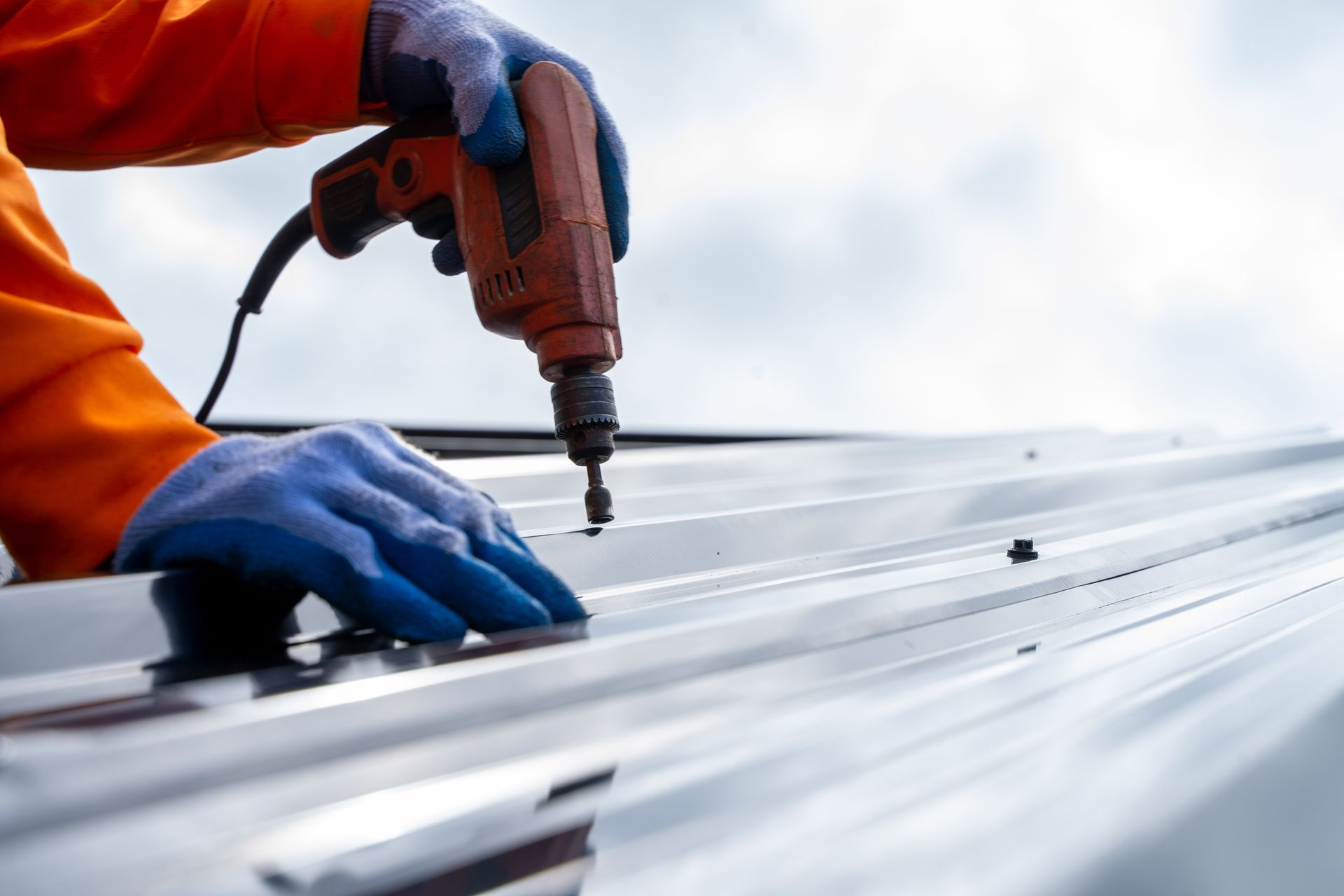 Worker drilling metal roofing panel with orange power tool and blue gloves