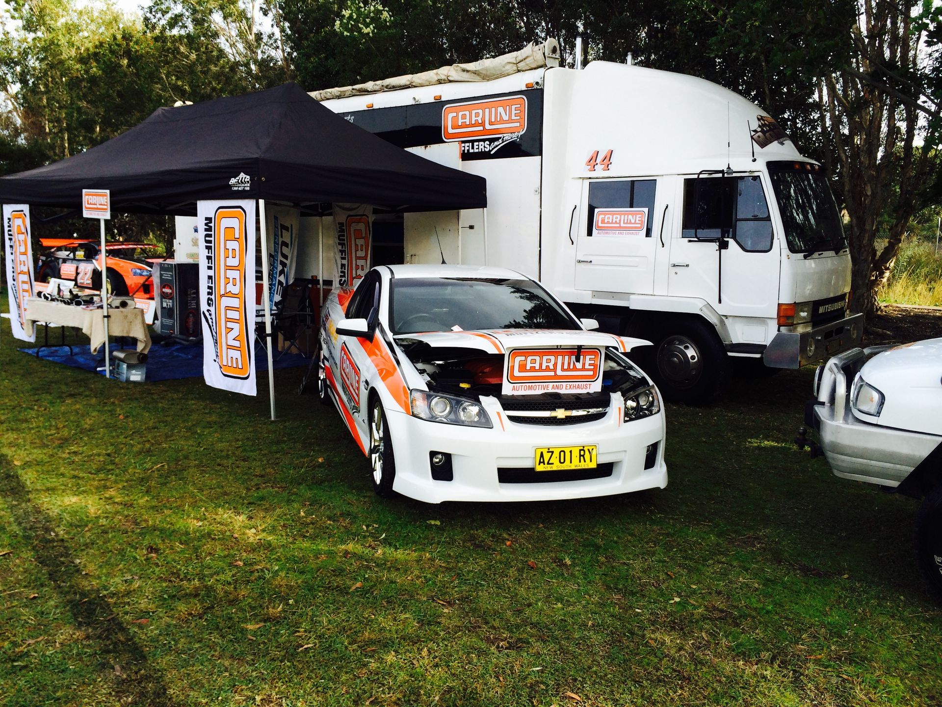 A white car with the hood open in front of a truck and tent setup for a car event on a grassy field.