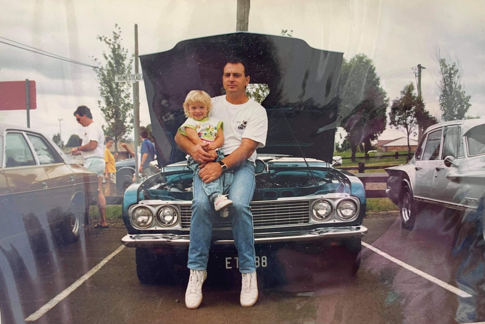 Man holding child in front of a blue car with hood open at an outdoor event.