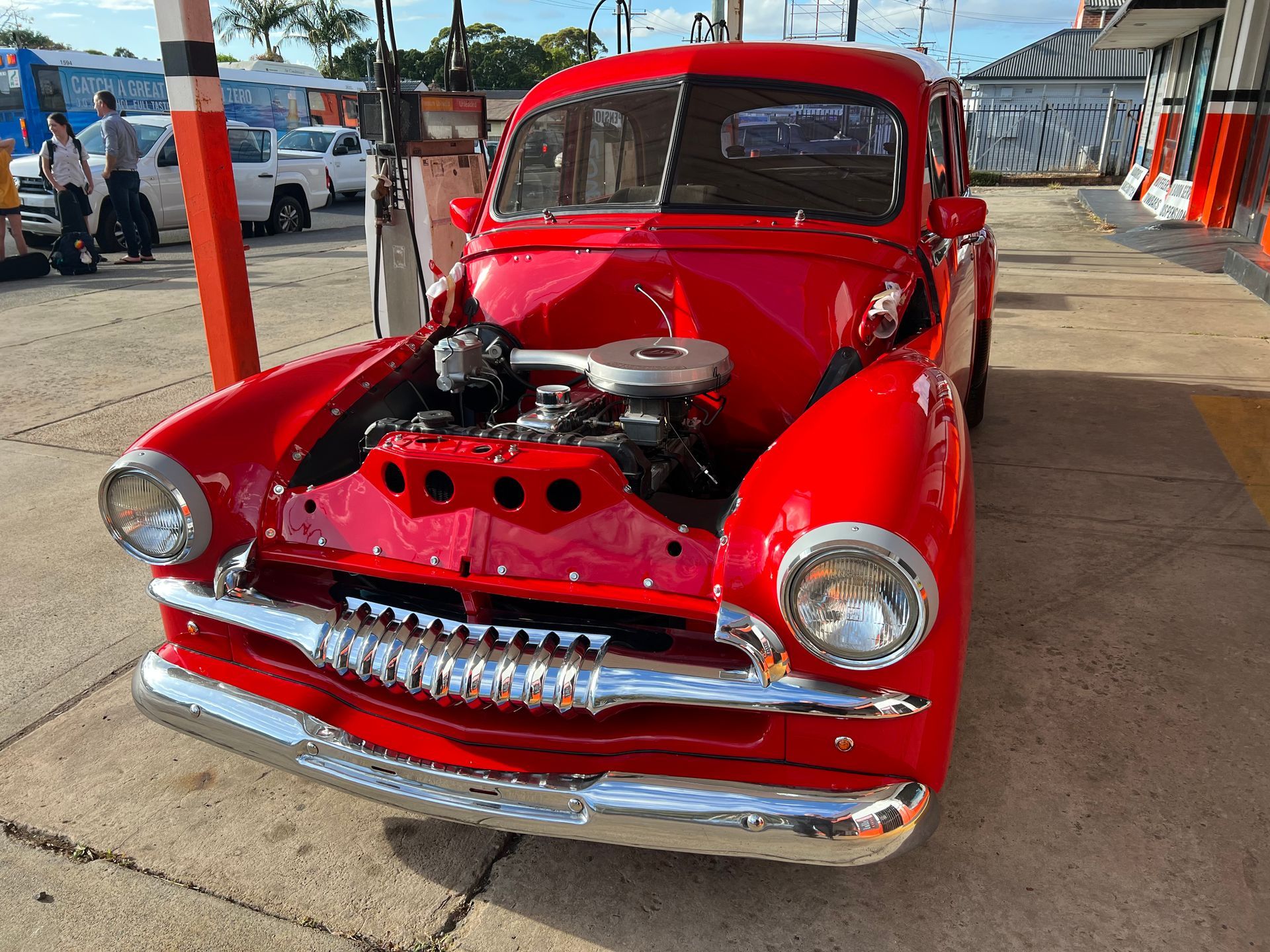 Red vintage car with open hood at a gas station, chrome details, sunny day.
