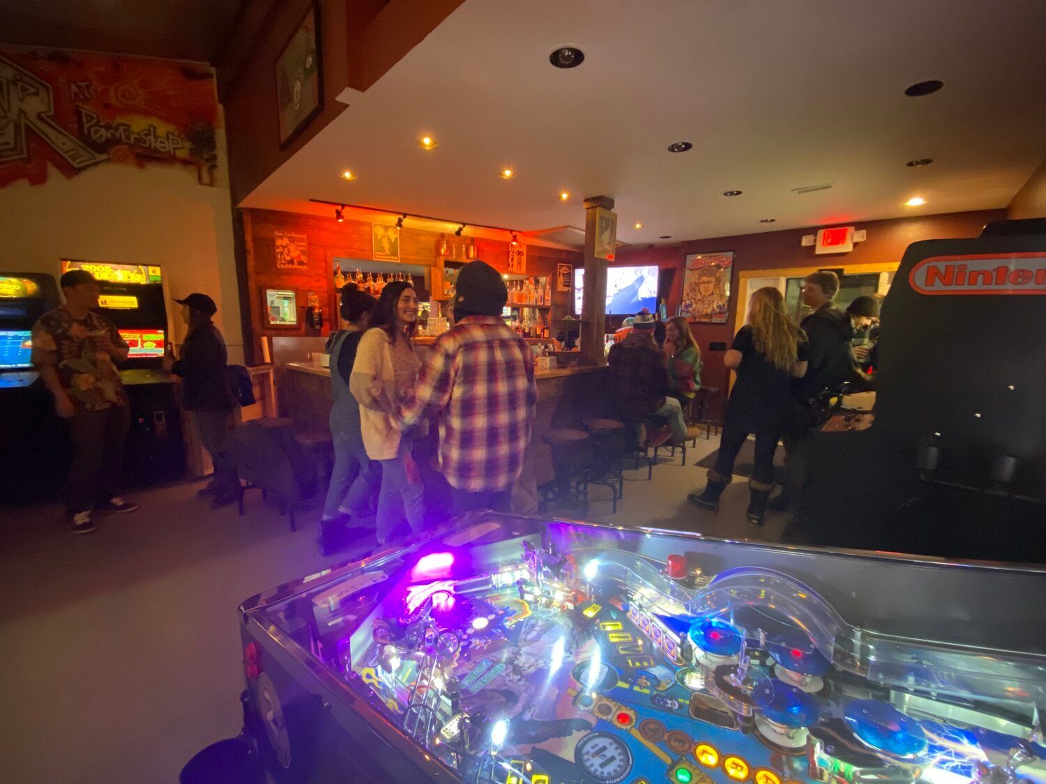 A group of people are playing pinball in an arcade