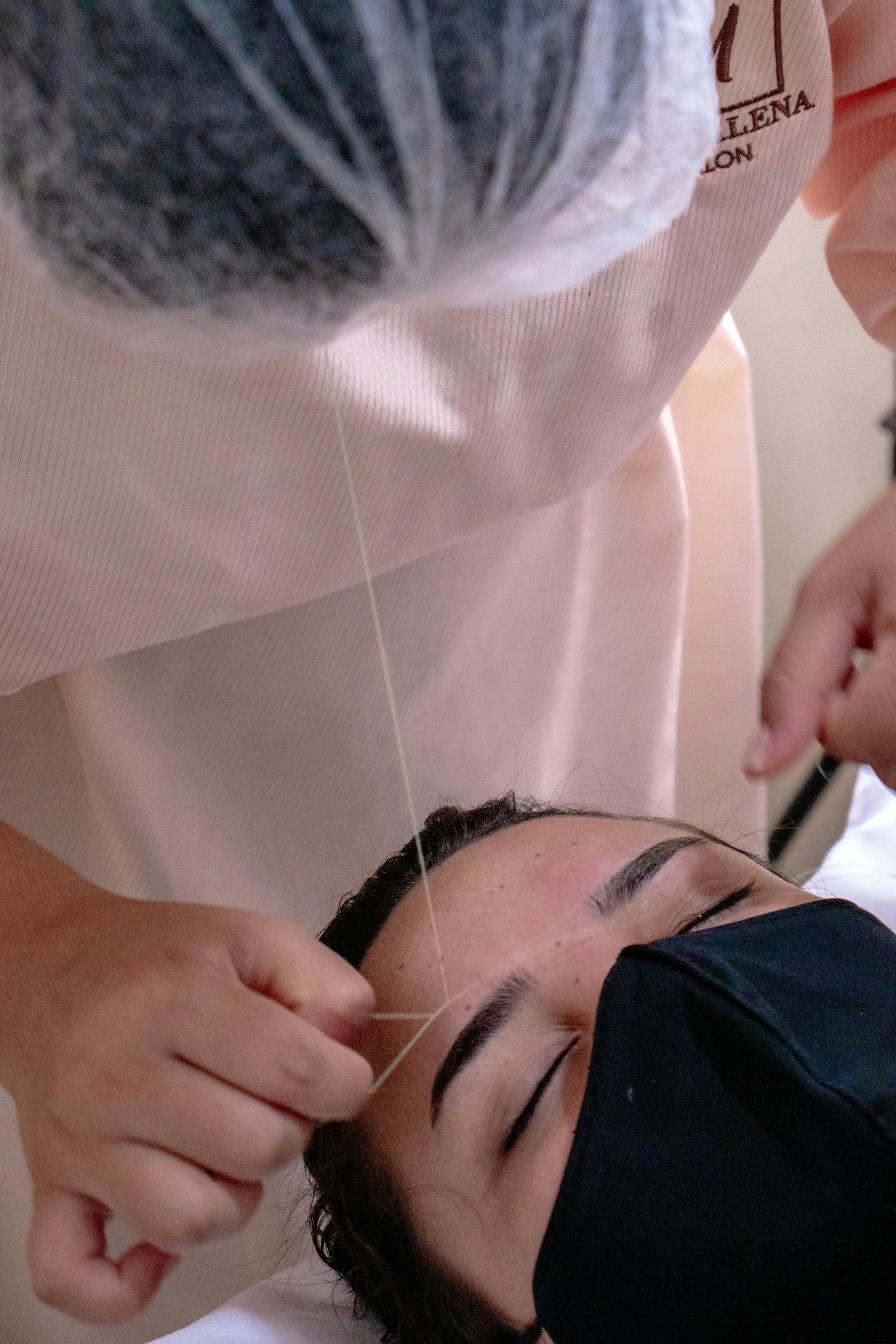 Person getting eyebrows threaded, face covered by mask, by a person with a white hair net.